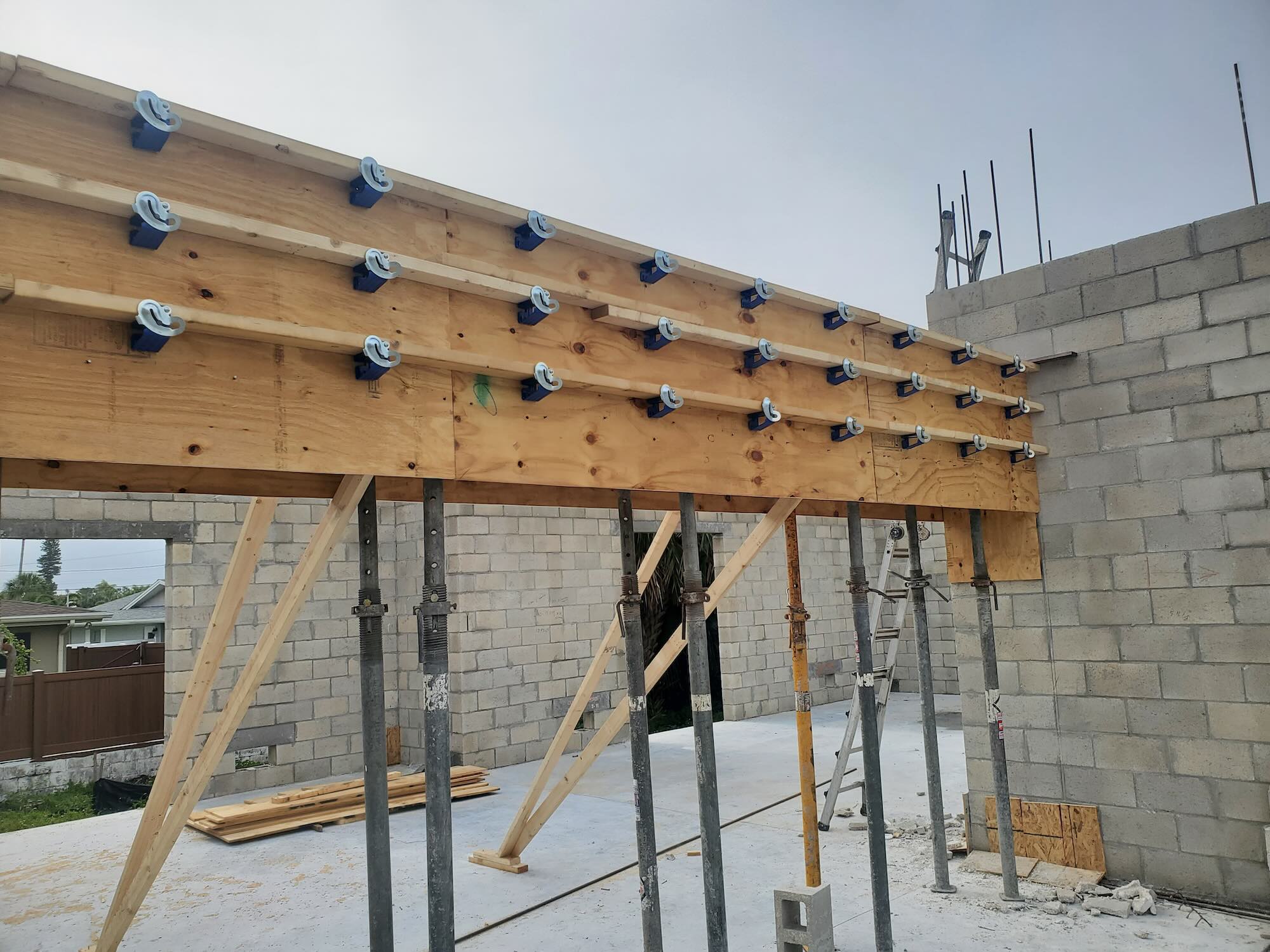 Close-up of a large built-up beam along a CMU wall, fitted with roughly two dozen blue joist hangers, shored from below on steel posts