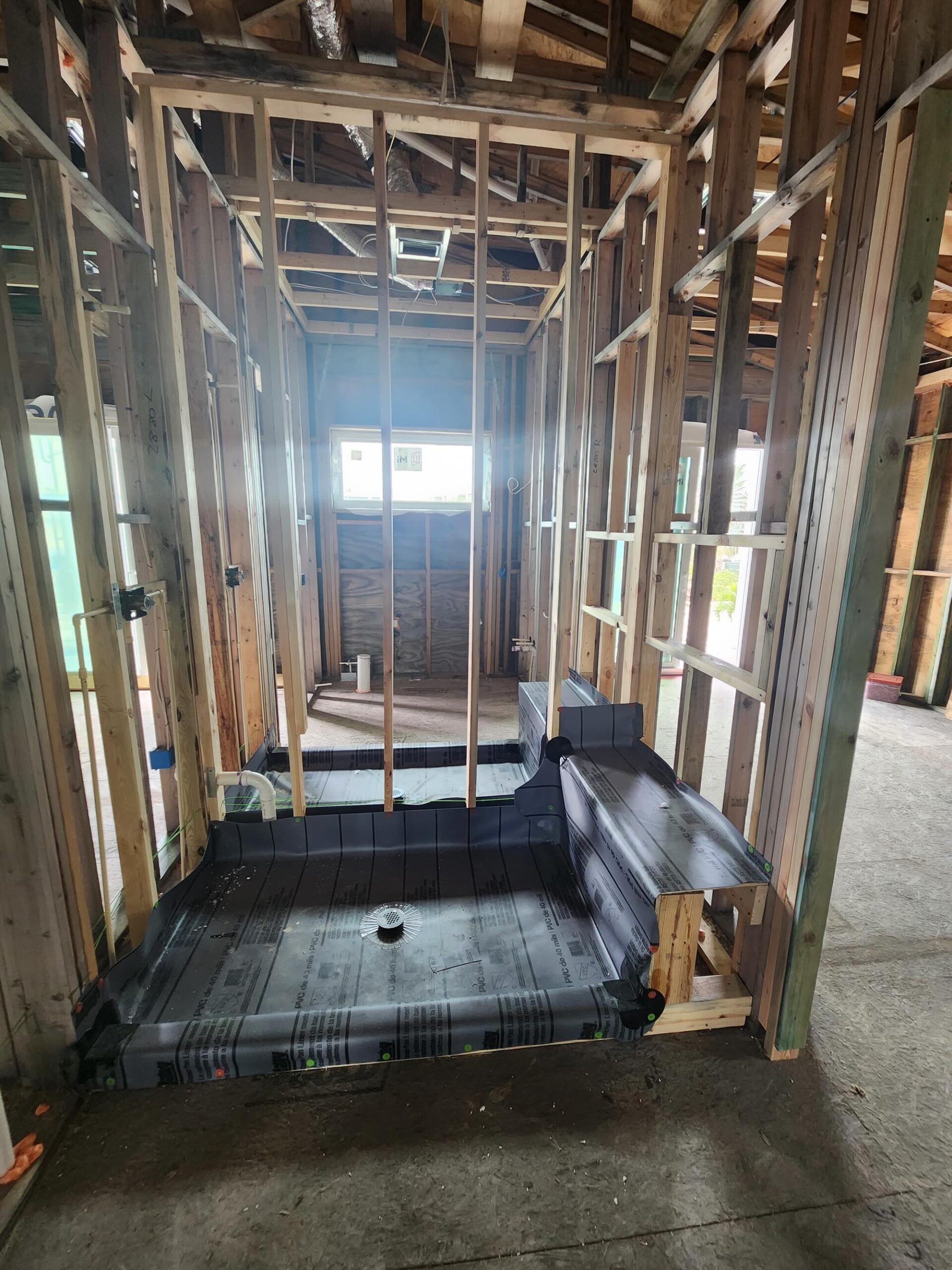 Interior wood-framed shaft cut into an existing house structure, with a PVC waterproof shower-pan style liner and center drain installed at the base, plumbing stubs visible in the surrounding walls