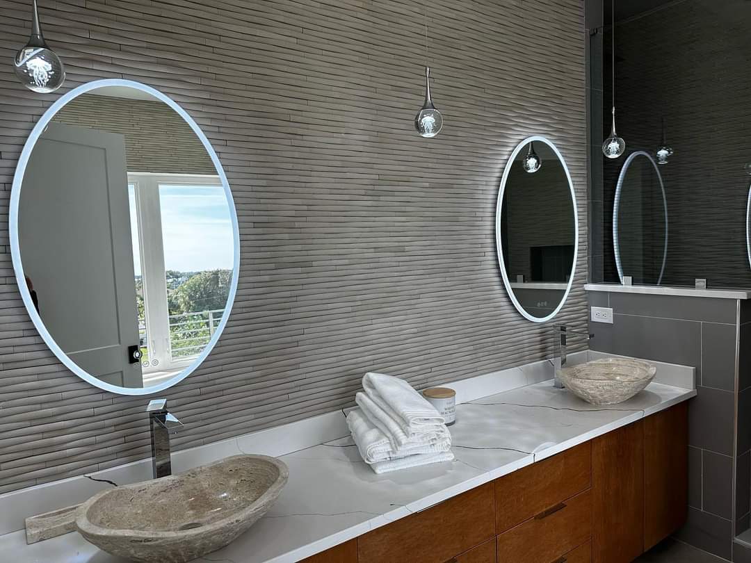 Primary bathroom with dual vessel stone sinks, backlit oval mirrors, and ribbed stone accent wall