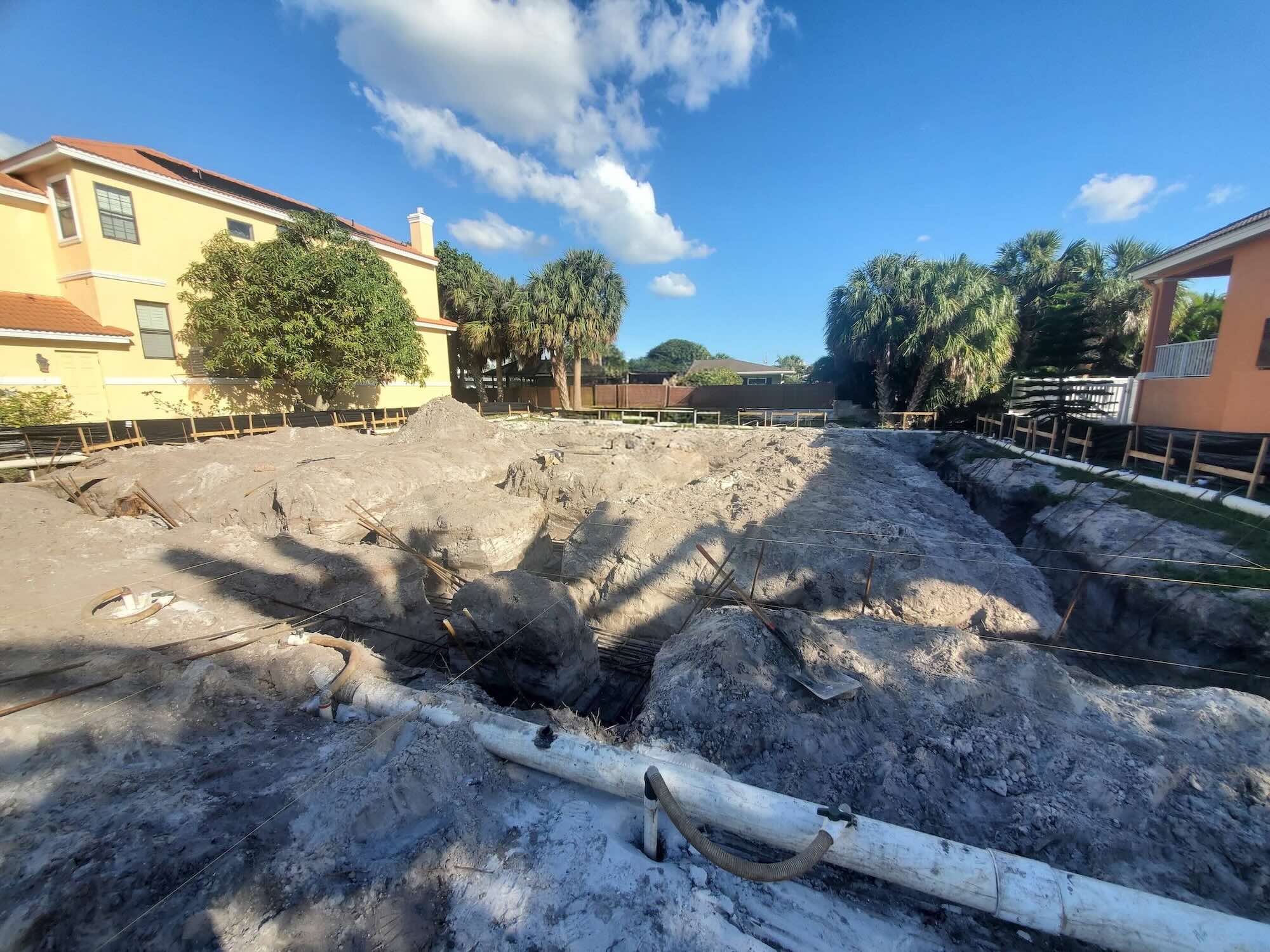 Wide shot of open footing trenches with rebar cages and vertical dowels tied in, ahead of a concrete pour, with neighboring houses visible beyond the lot line