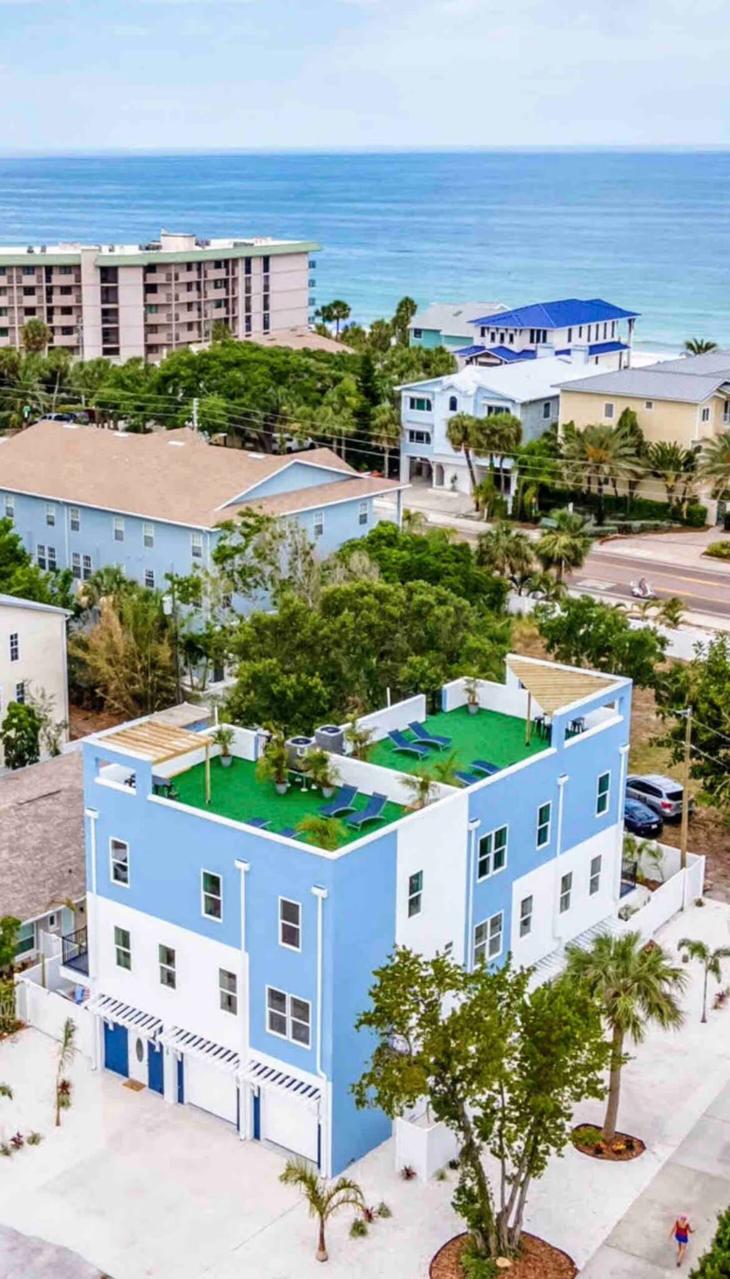 Aerial view of a blue and white three-story coastal home with a rooftop deck, palm trees, and the ocean visible beyond