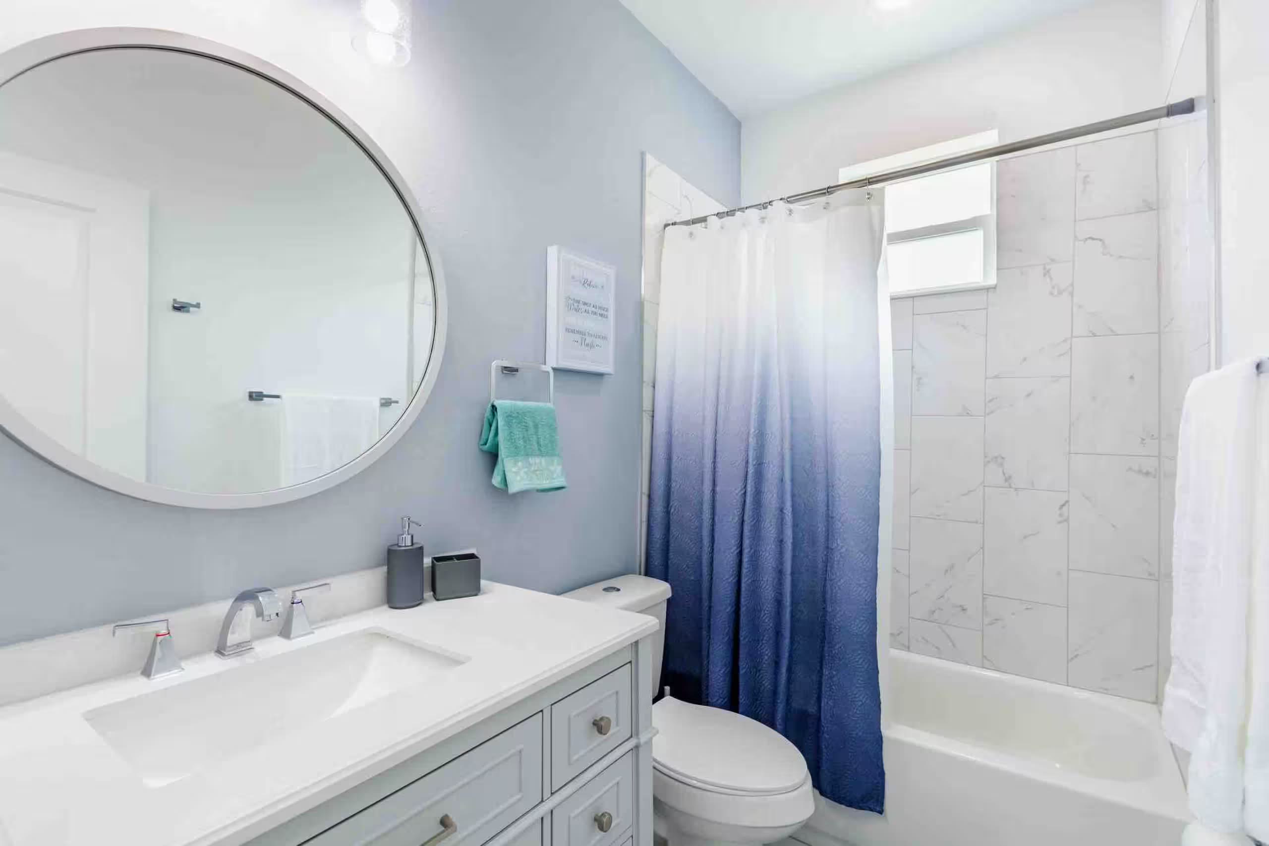 Bathroom with a round mirror, white vanity, and a tub/shower combo behind a blue ombre curtain in a marble-tiled surround