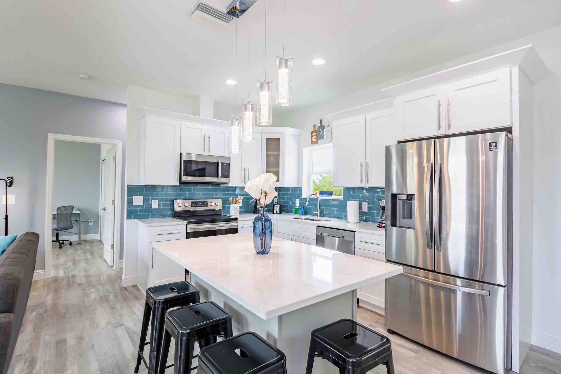 White kitchen with a waterfall-edge island, blue subway tile backsplash, black barstools, and stainless steel appliances
