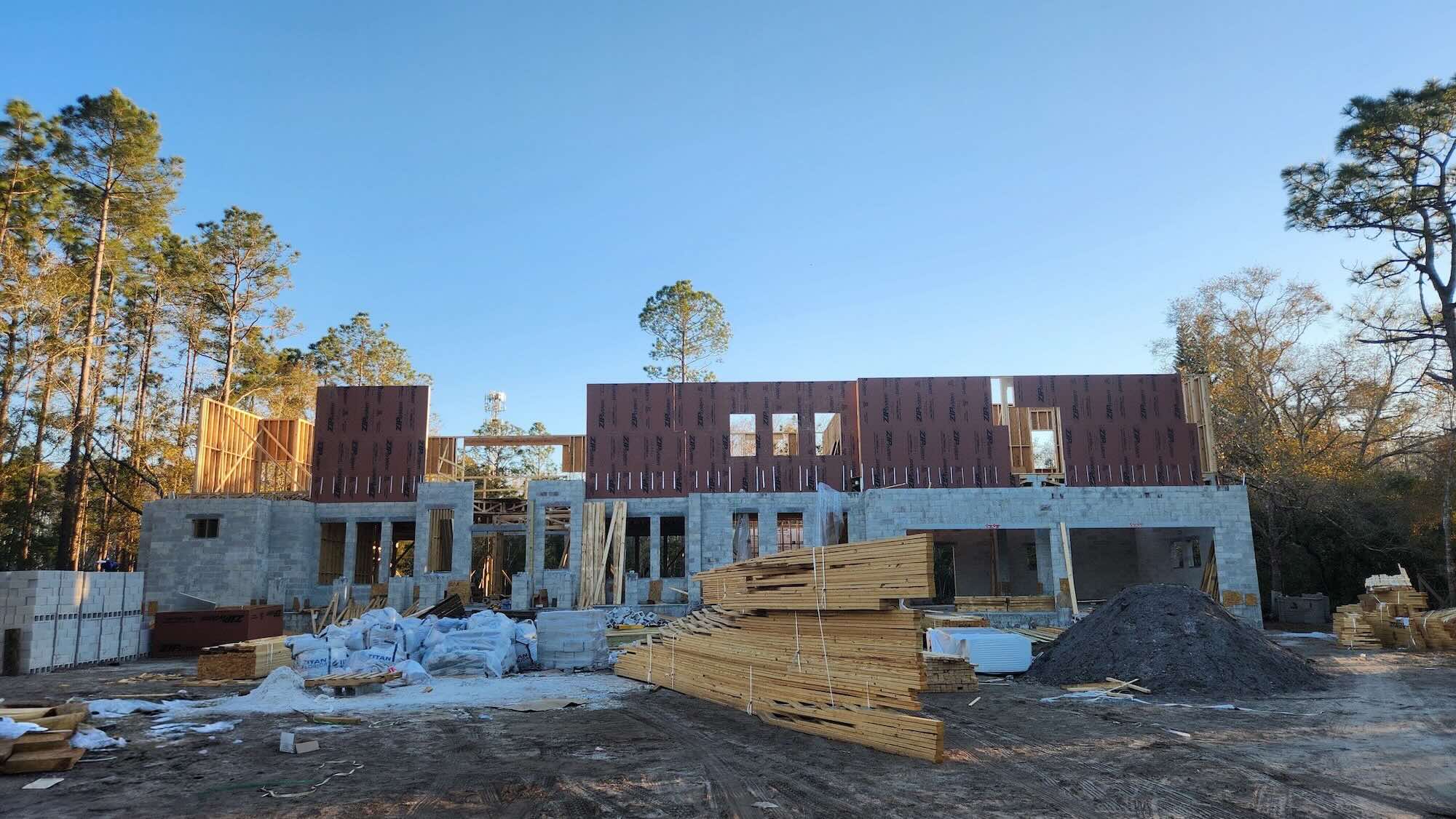 Two-story structure under construction at dusk, with finished concrete block walls on the first floor and wood-framed second-floor walls wrapped in ZIP System sheathing above, surrounded by stacked lumber and building materials