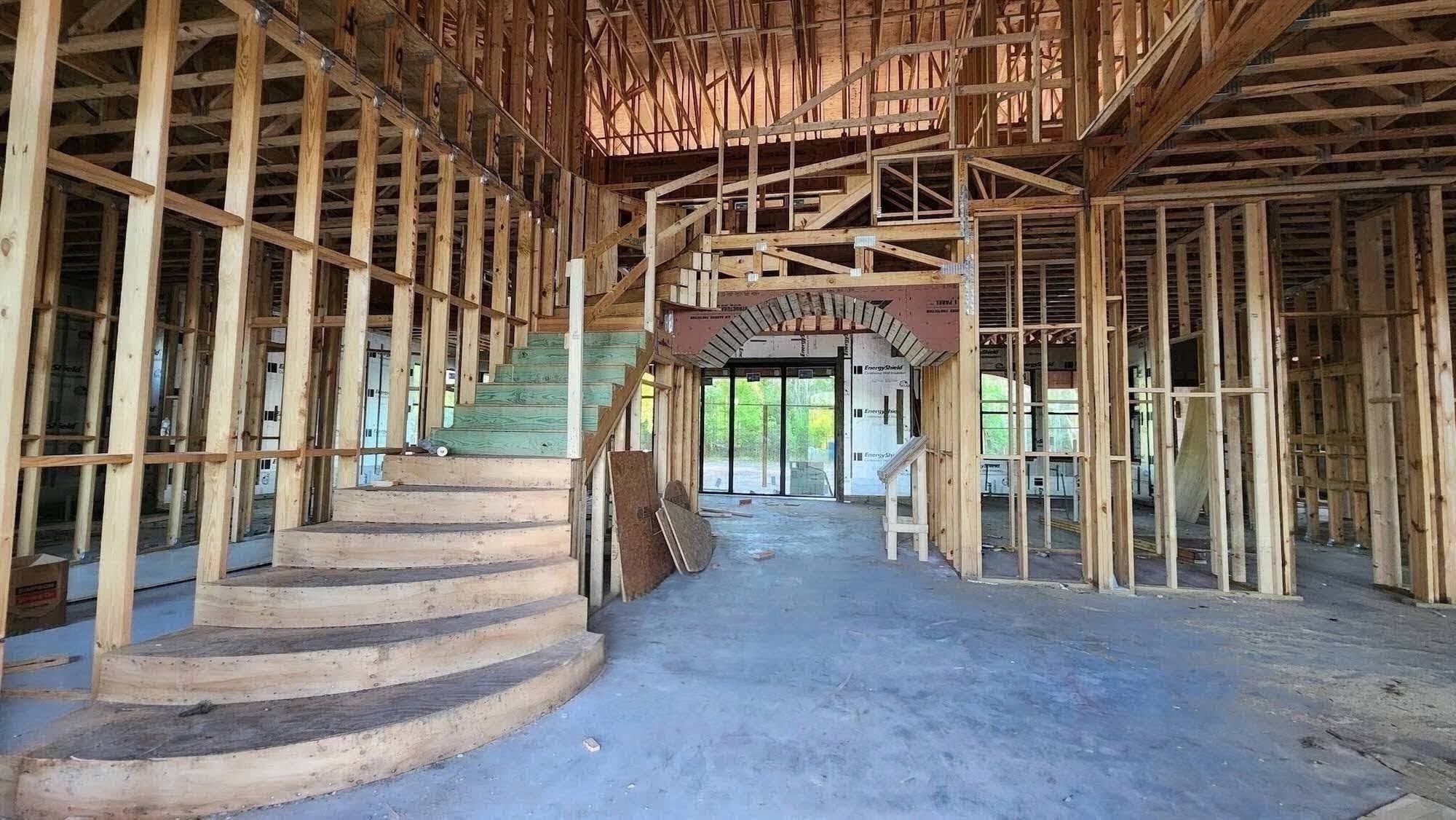 Interior of a framed home under construction showing a curved staircase with green-treated lower steps and natural wood upper steps, rising beneath a tall vaulted ceiling of exposed roof trusses, with an arched wood-framed opening leading to sliding glass doors at the rear