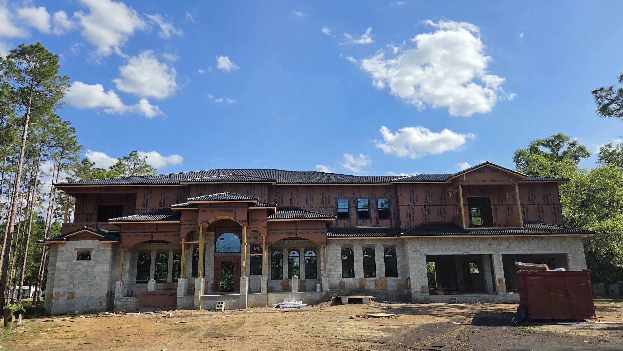Two-story home under construction with a completed dark tile roof, ZIP System sheathing on the upper-floor exterior walls awaiting cladding, arched window and door openings in the concrete block lower level, and a rental dumpster in the front yard