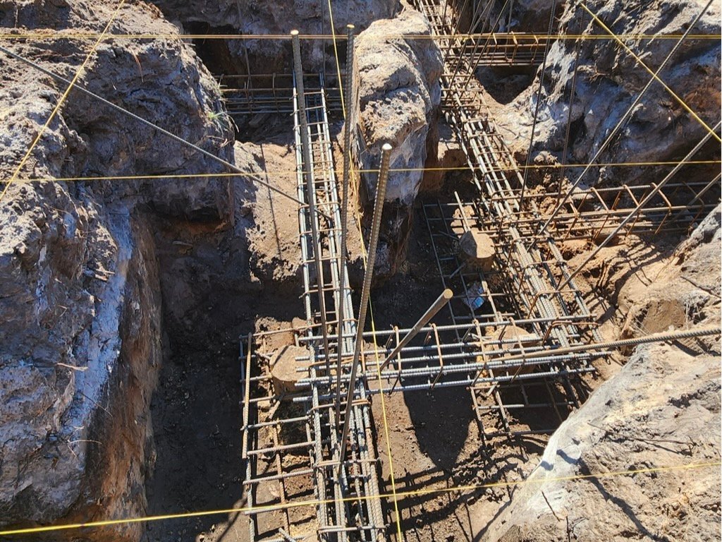 Overhead view of a steel rebar cage set into an excavated footing trench, with intersecting reinforcement grids and string lines marking the layout above