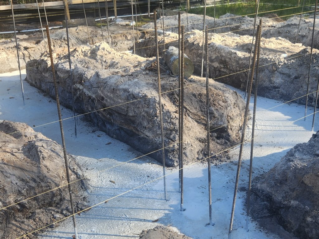 Excavated footing trenches for a home's foundation, with vertical rebar dowels and string layout lines running along both sides of a corner, sandy excavated soil piled at the trench edges.