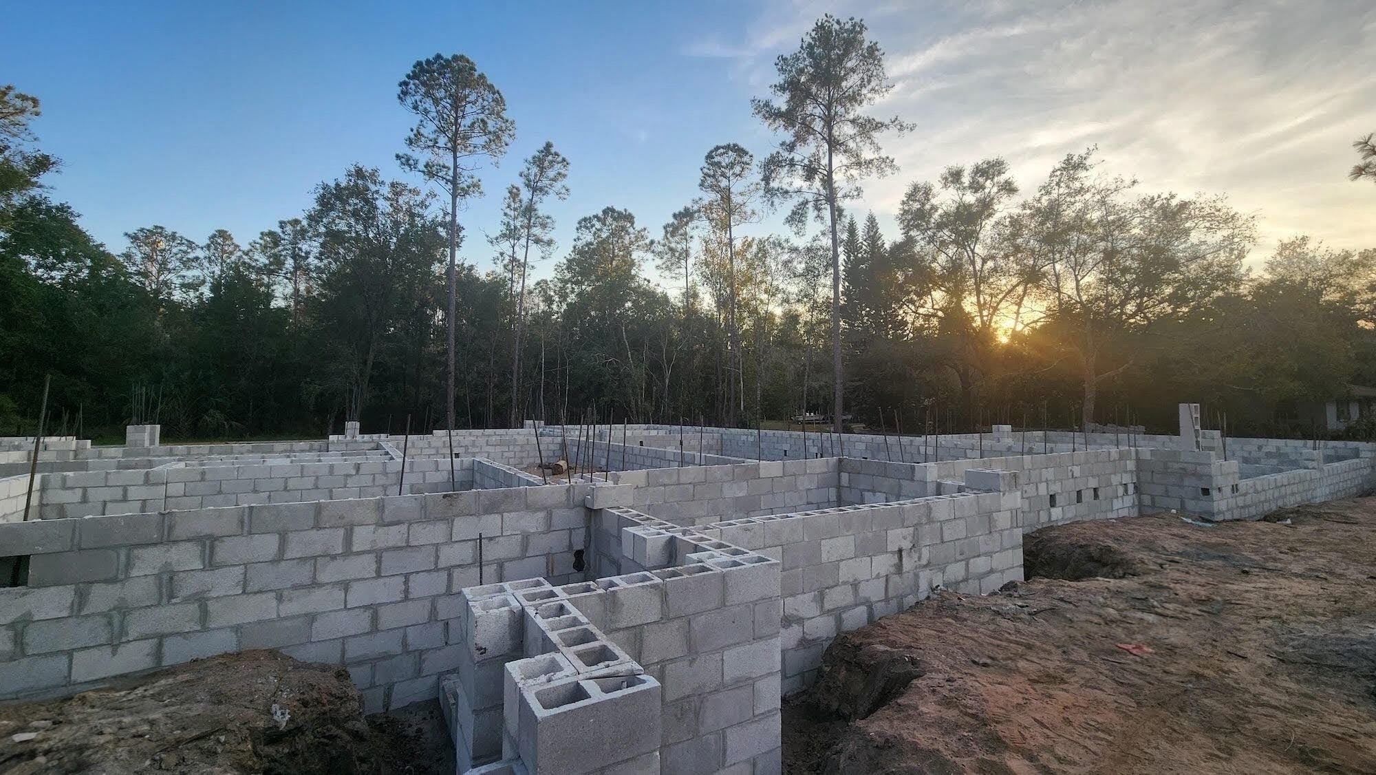 Low concrete block foundation walls outline the footprint of a home under construction, with vertical rebar dowels exposed at wall intersections and the sun setting through trees in the background