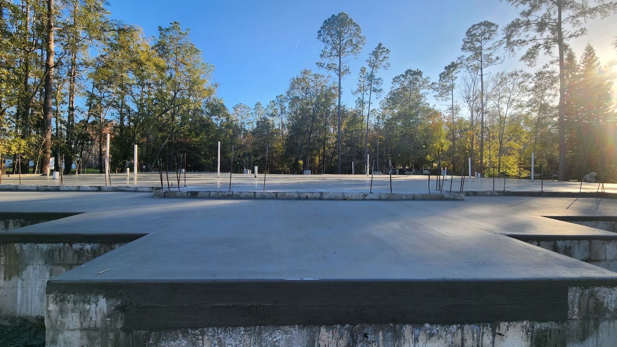 A worker uses a walk-behind power trowel to finish a freshly poured concrete slab, with PVC plumbing stub-outs and rebar dowels protruding from the surface and trees lining the property in the background