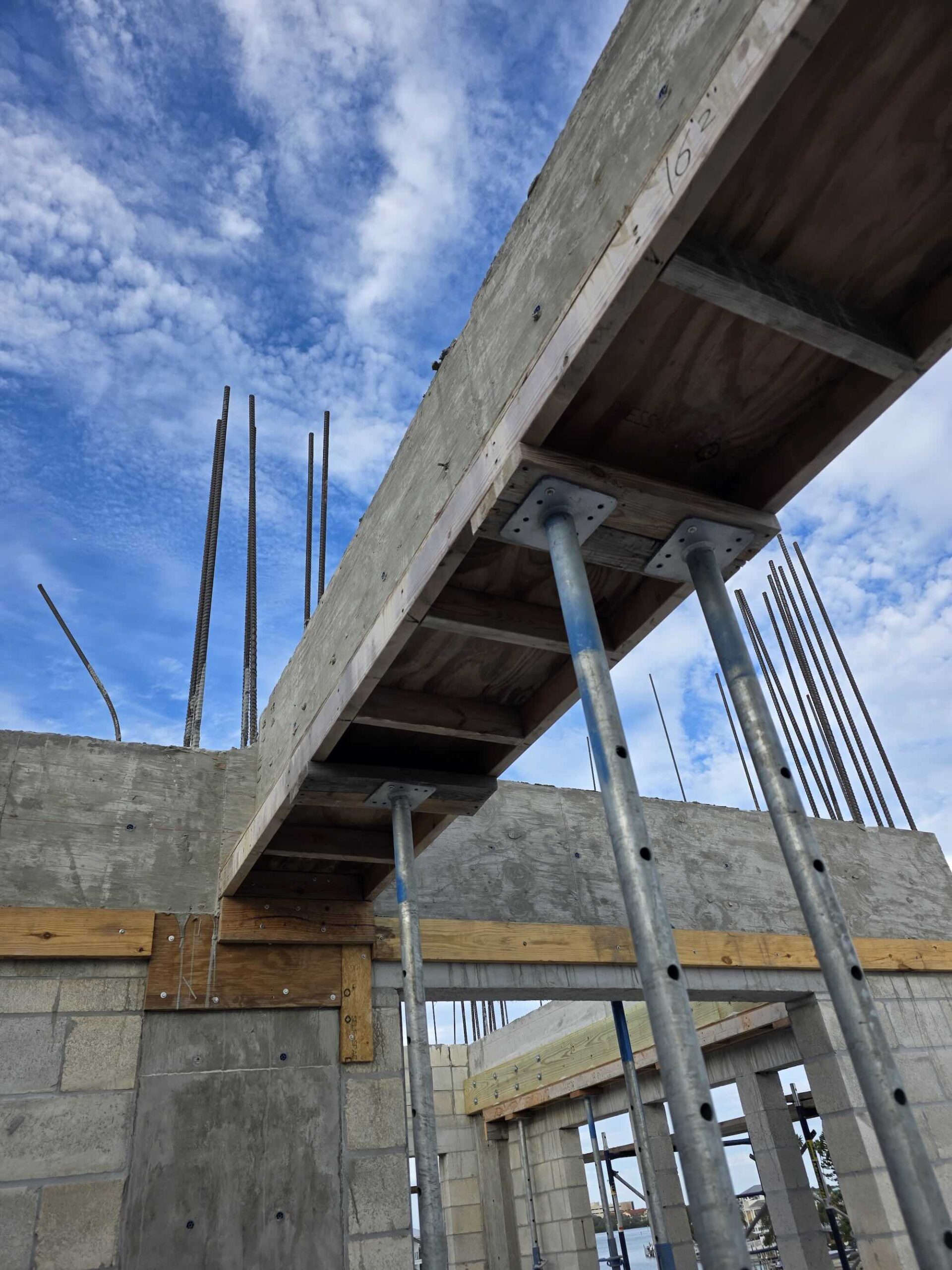 Underside view of a concrete beam formed in wood with steel shoring posts and protruding rebar