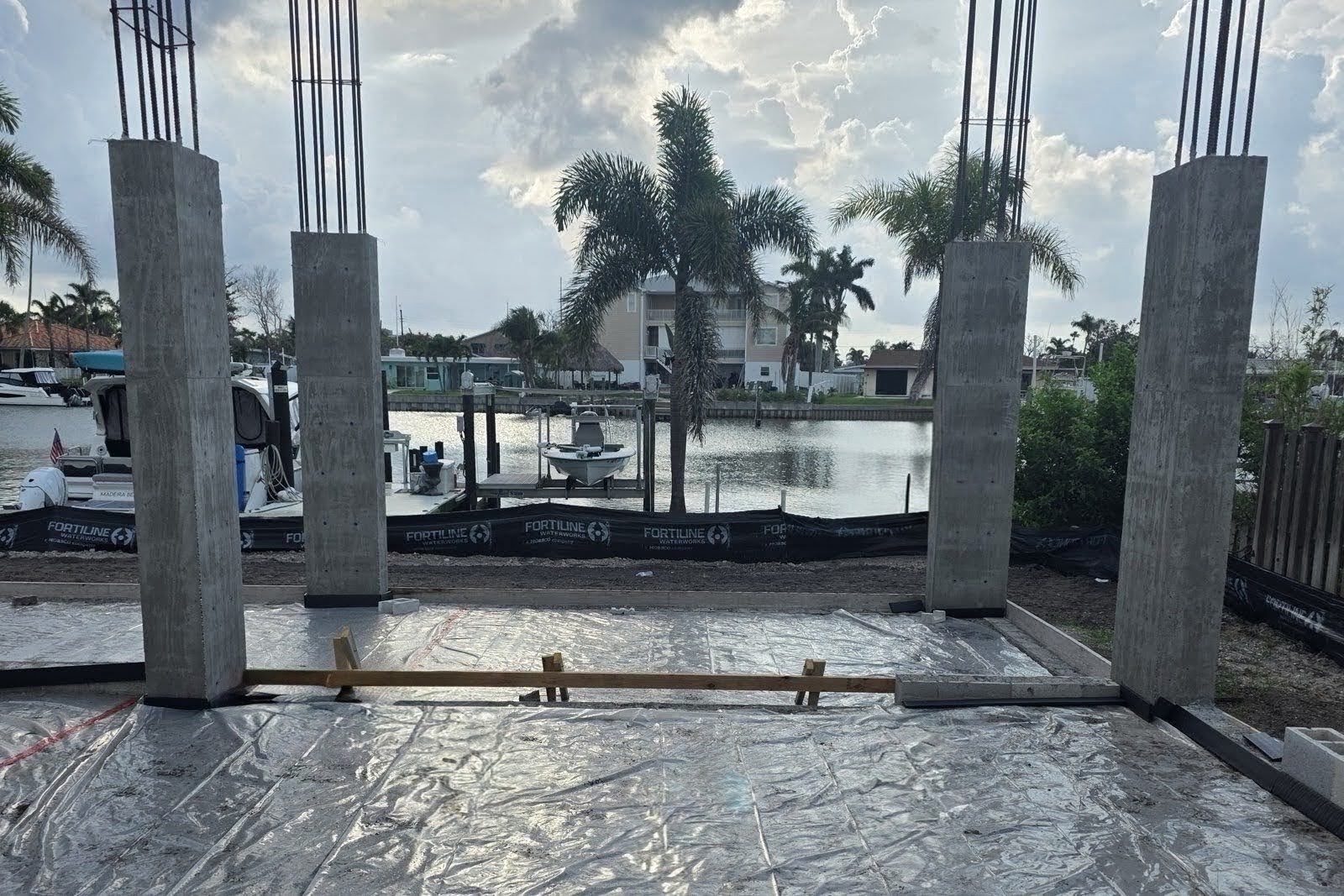 Concrete columns with protruding rebar rising from a plastic-sheeted ground slab at a waterfront construction site