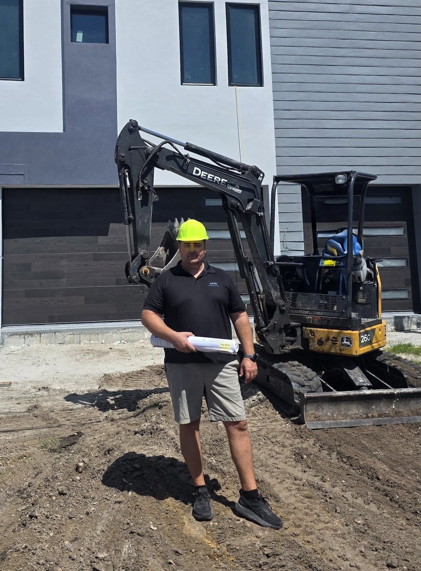 Sasa Nikic, owner of Nikic Construction, standing on a job site holding rolled plans in front of a mini excavator