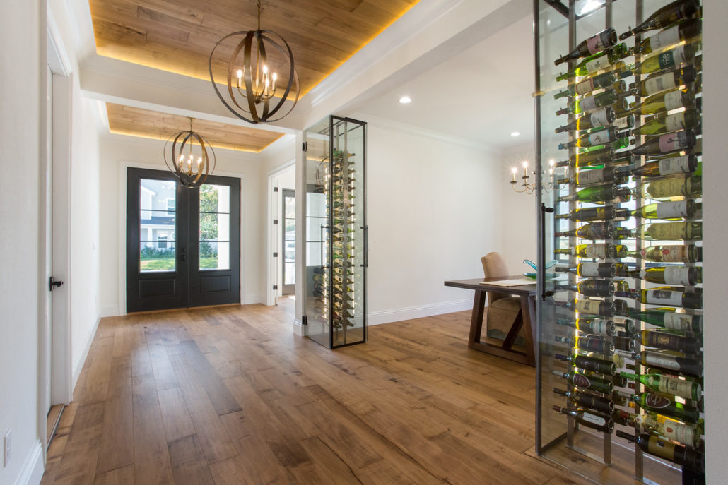Glass-enclosed wine display wall beside a wood dining table, under a wood-paneled ceiling with cove lighting