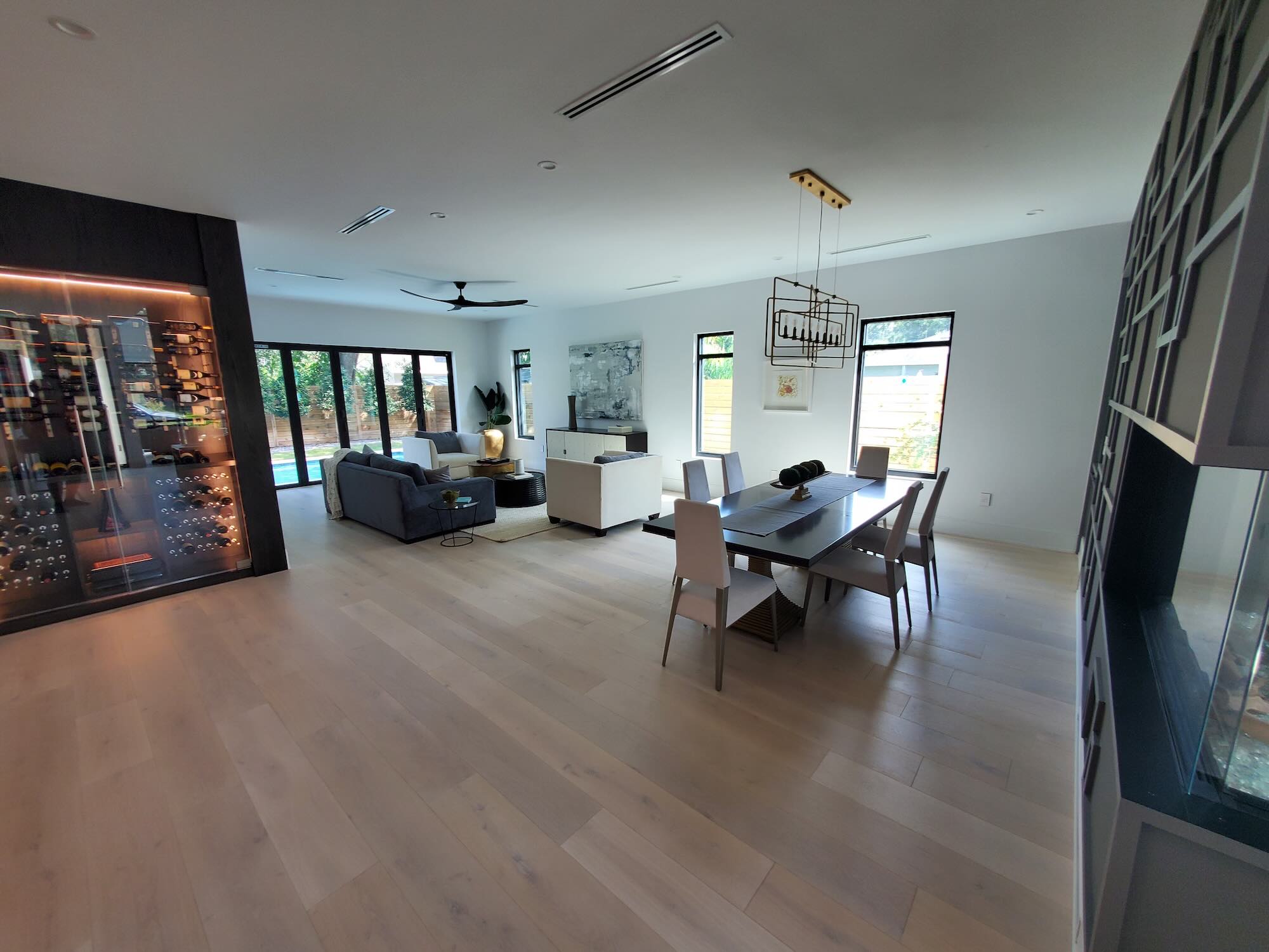 Dining and living room with a glass-enclosed wine wall in the foreground and a folding glass door open to a pool visible beyond