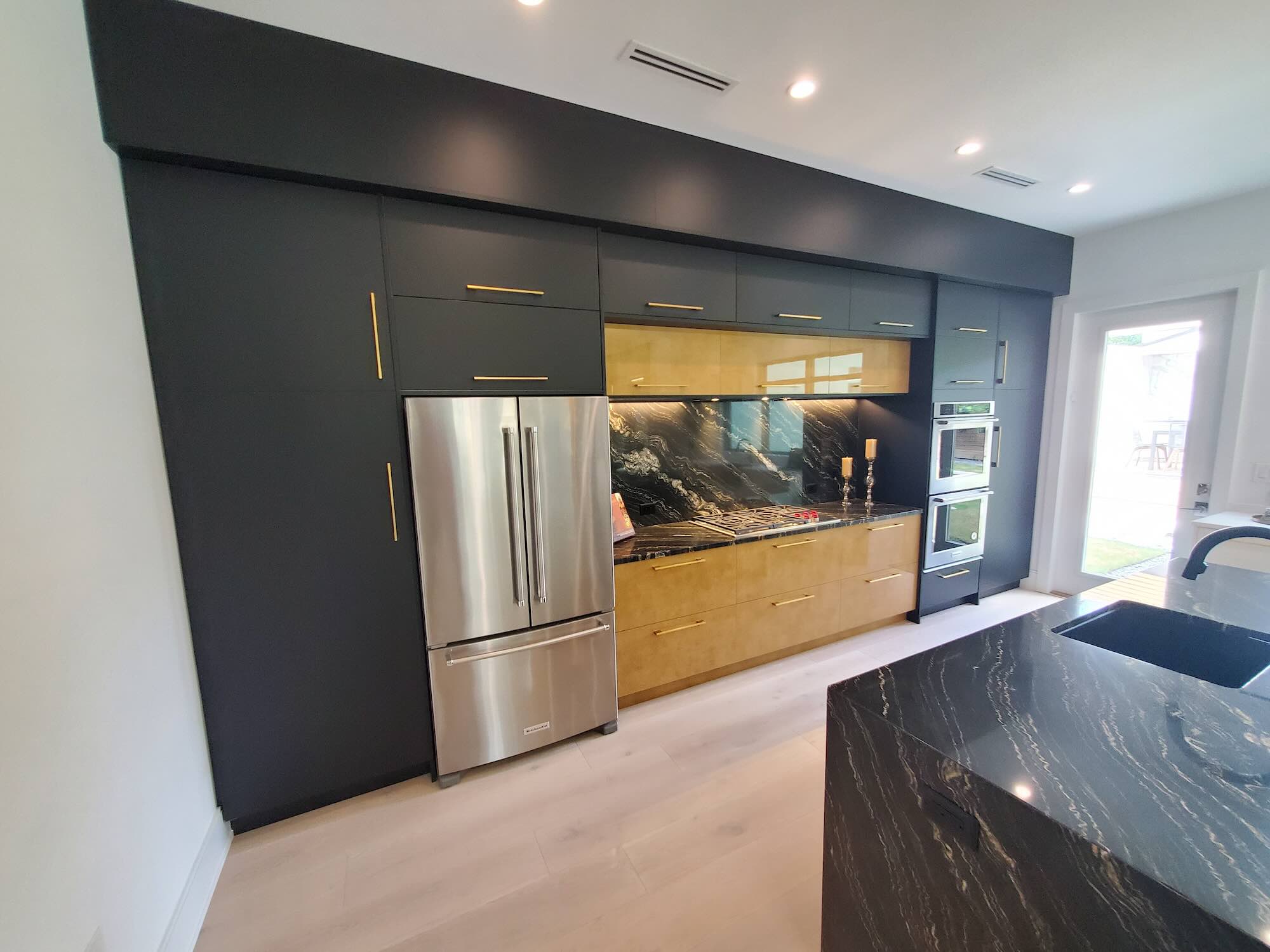 Kitchen wall with charcoal cabinetry, a light wood cabinet run, full-slab black marble backsplash, and a built-in stainless refrigerator and double ovens