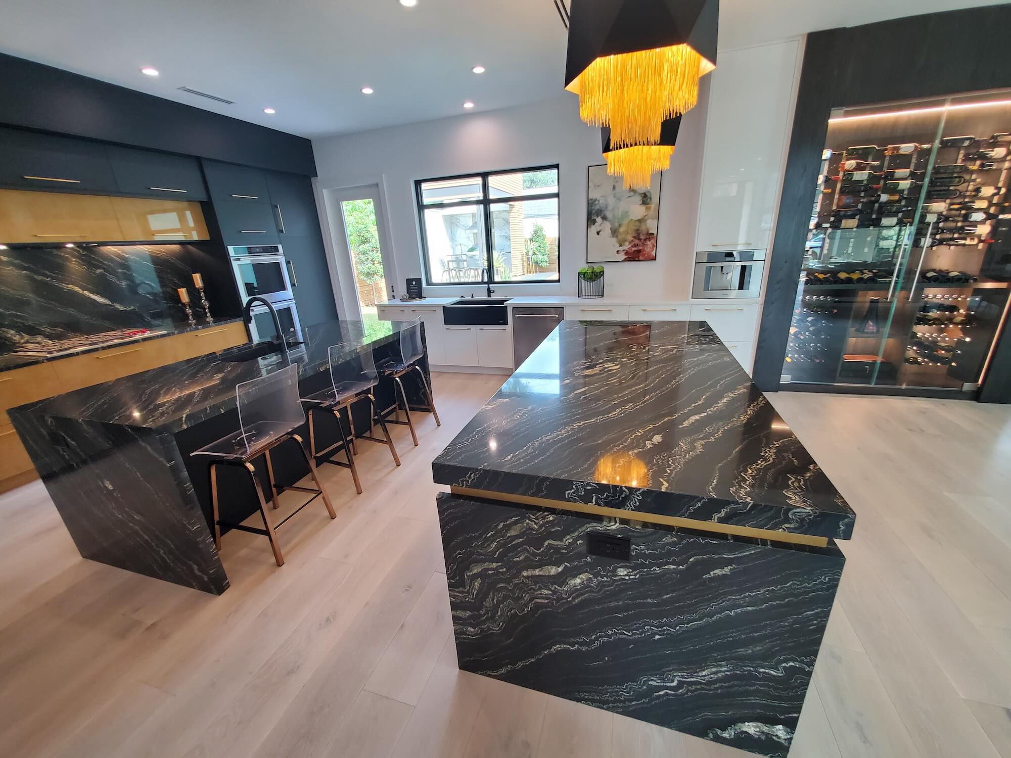 Kitchen island with black stone waterfall countertop, clear acrylic barstools, a faceted gold pendant light, and a glass wine wall in the background