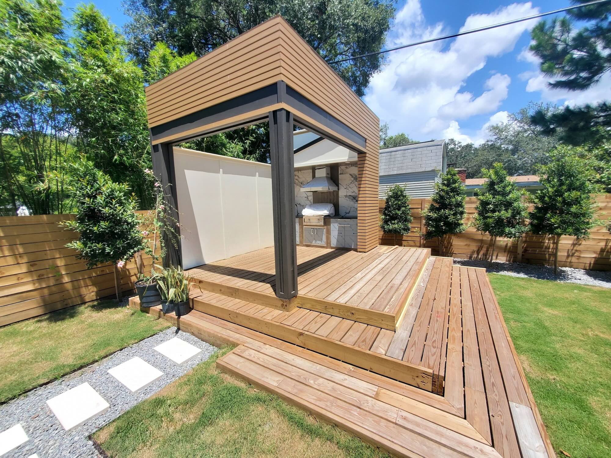 Outdoor cabana with a wood slat roof and a built-in outdoor kitchen with a marble backsplash and stainless grill, set on a wood deck