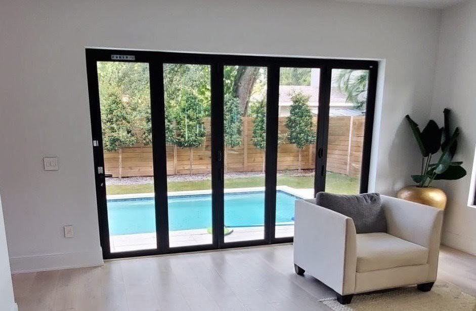 Living room with a folding glass wall opening onto a pool, wood privacy fence, and young trees along the back property line