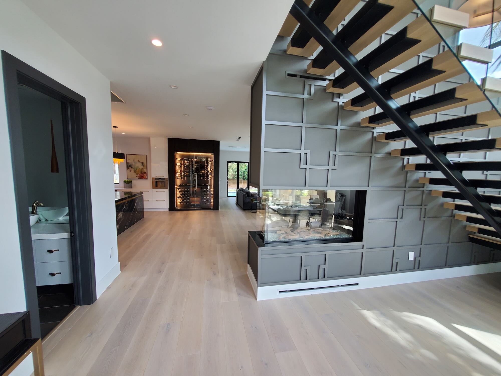 Floating wood staircase with black steel stringers beside a three-sided glass fireplace and a lit glass wine wall down the hallway