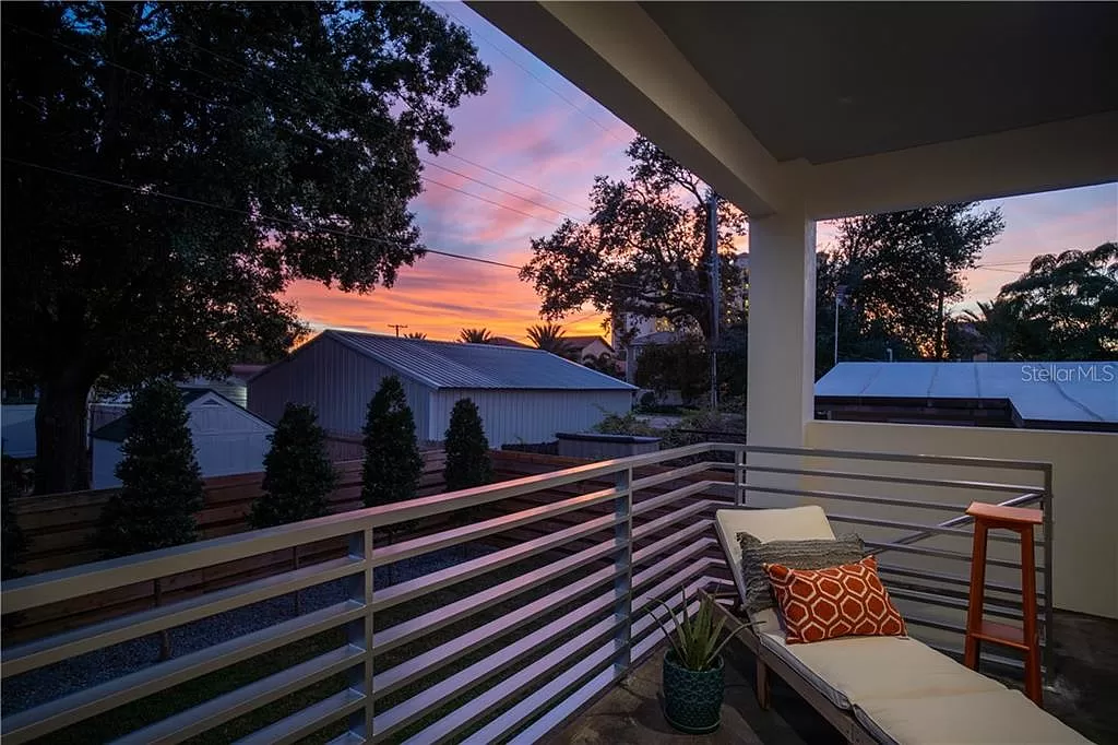 Covered balcony at sunset with a lounge chair and orange geometric-patterned cushions, overlooking mature trees and neighboring rooftops