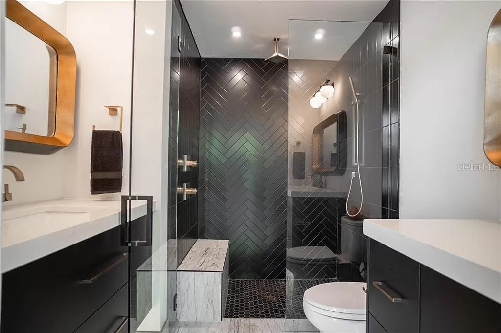 Bathroom with a walk-in shower finished in black herringbone tile, brass fixtures and mirror frame, and a two-sink vanity with white countertop