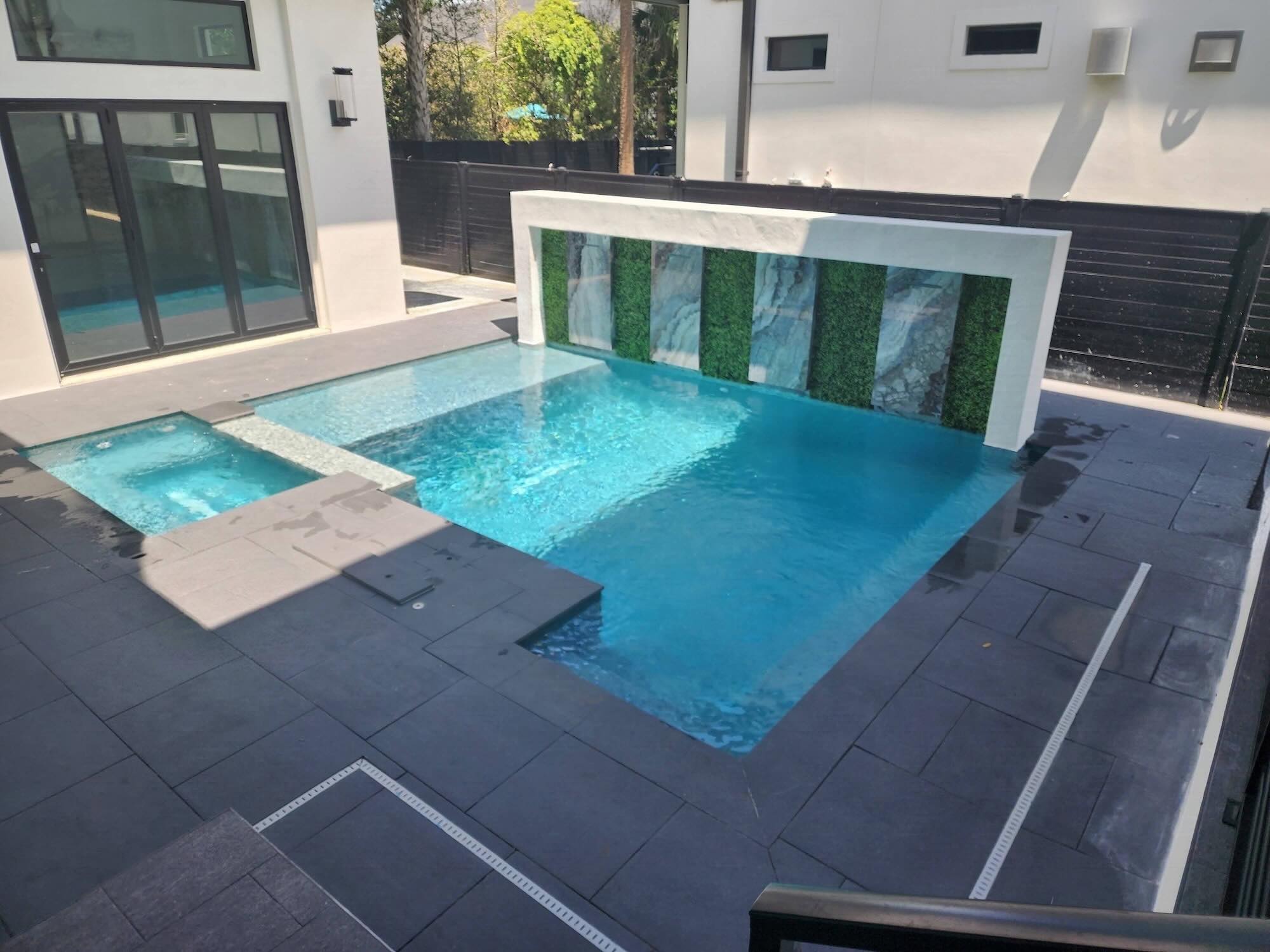 Rectangular pool and spa with stone decking and a vertical accent wall with greenery inlay, adjacent to the home's rear entry