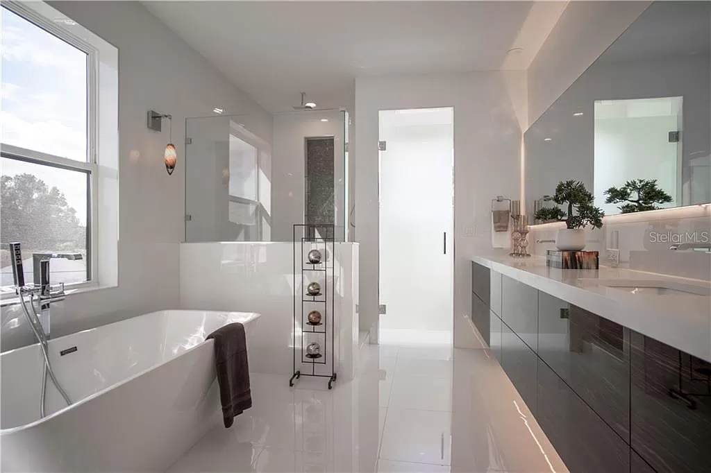 Primary bathroom with a freestanding soaking tub beneath a window, glass-enclosed shower, and a long dark wood vanity with backlit mirror