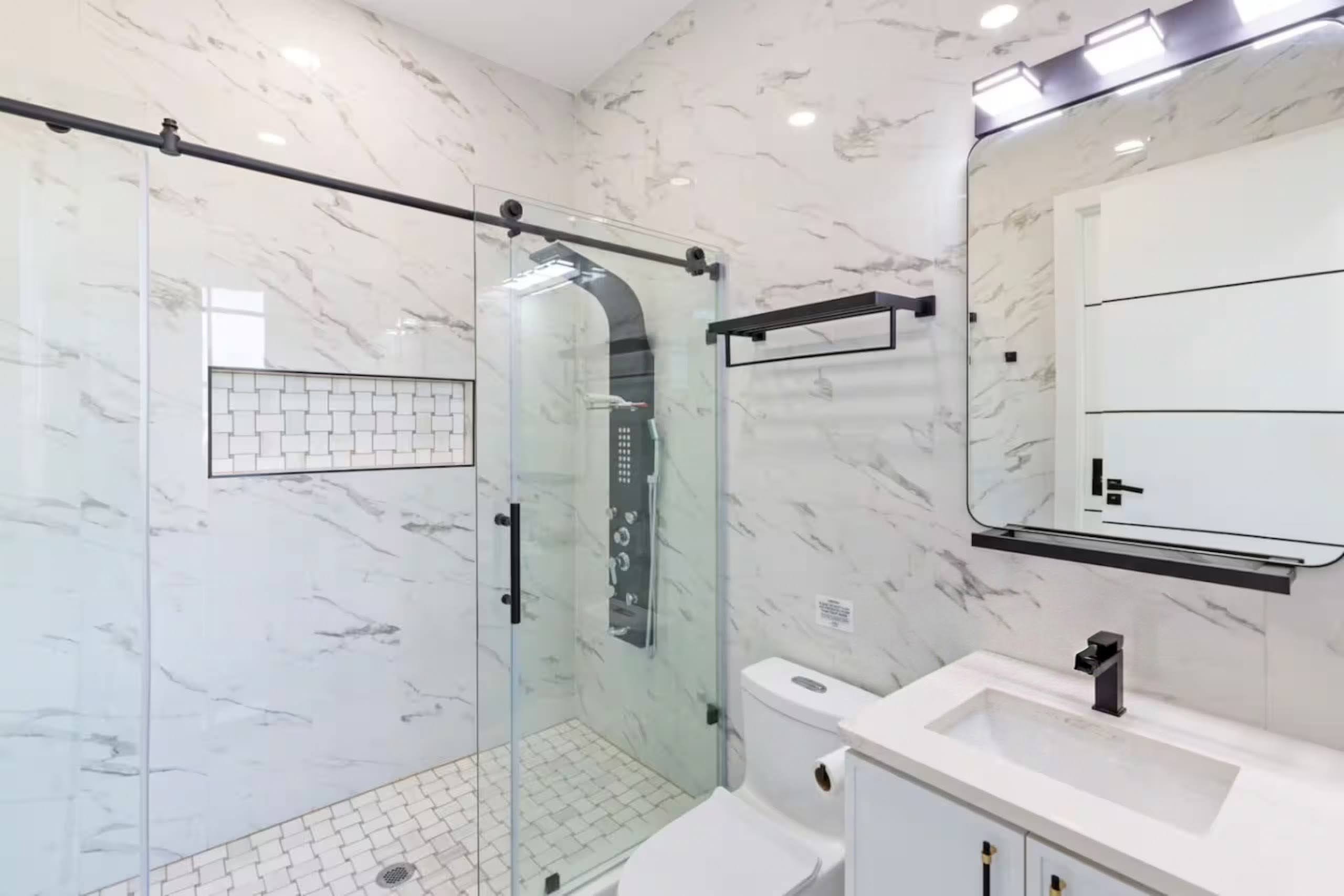 Marble bathroom with a barn-door glass shower enclosure, tiled niche shelf, freestanding vanity, and backlit mirror