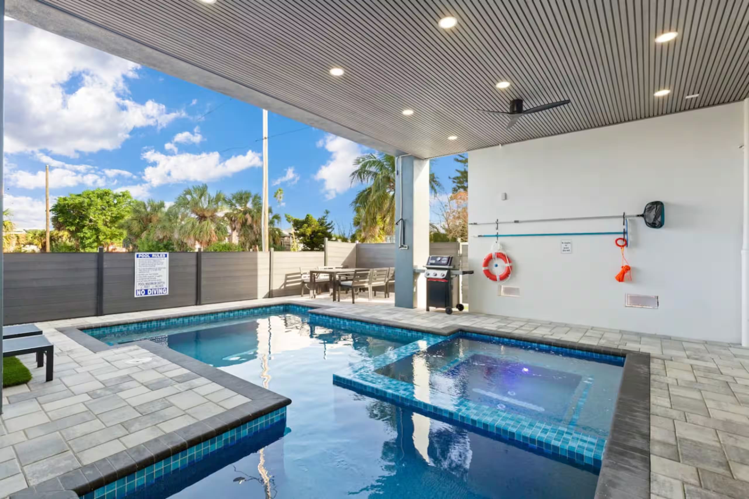 Covered ground-floor pool and spa with gray tile decking, built-in grill, and palm trees visible beyond the fence
