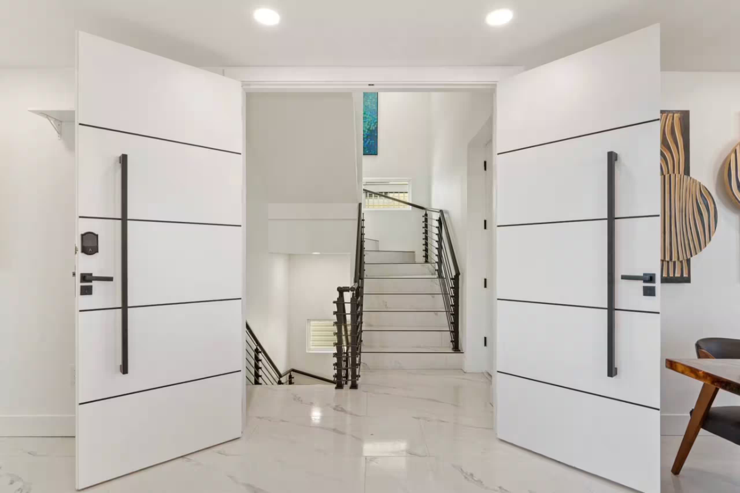 Oversized white double-panel entry doors opened onto a marble-floored foyer with a floating staircase and black metal railing beyond