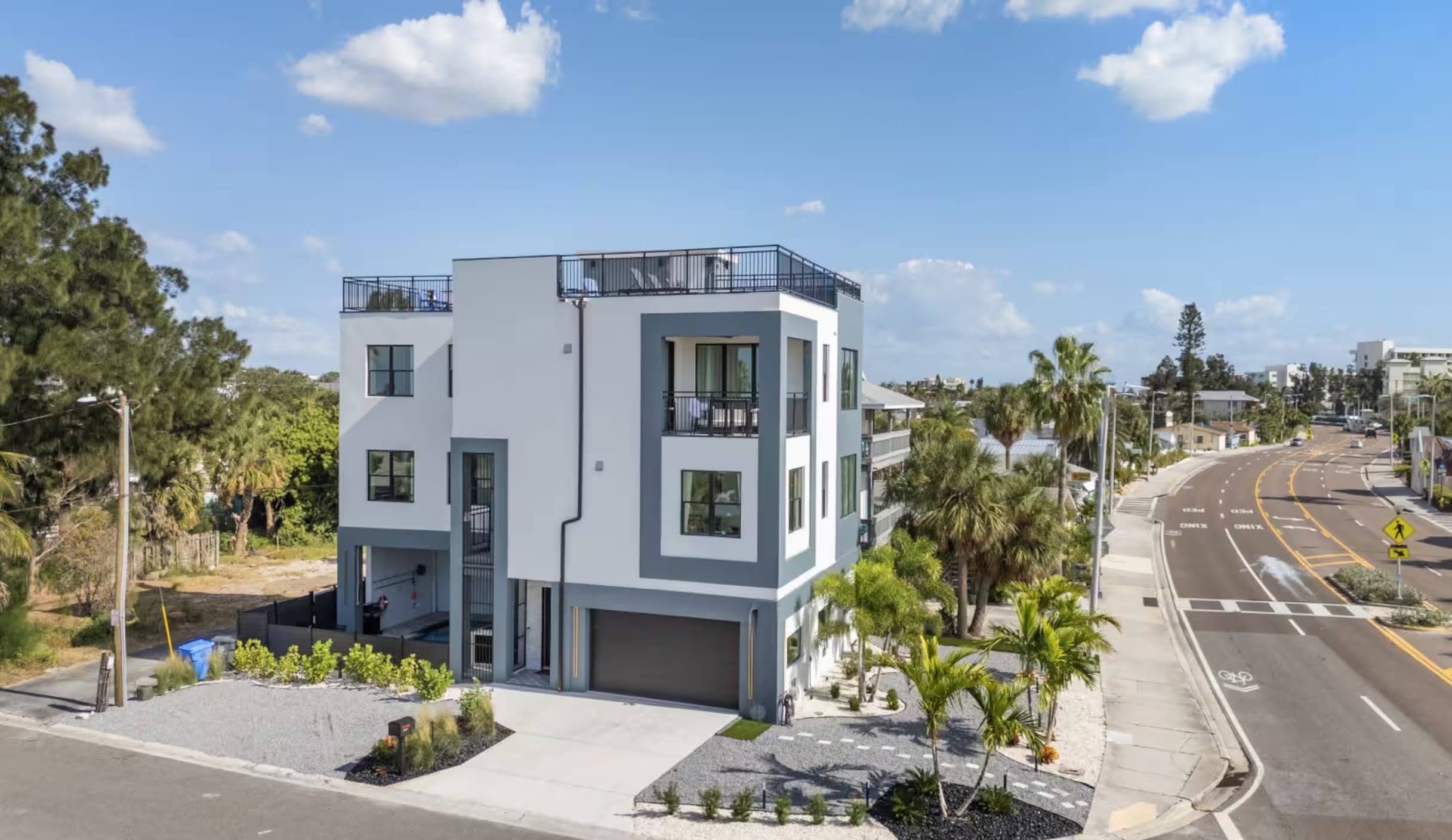 Three-story modern coastal home with white and dark gray stucco, black-framed windows, rooftop deck railing, and palm landscaping