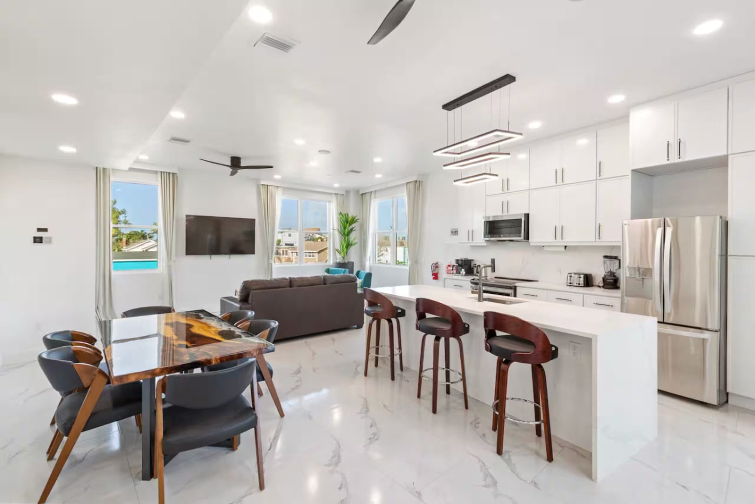 Open-concept kitchen with a white marble island, wood-and-leather bar stools, and stainless refrigerator, with the dining table and living room visible in the same room
