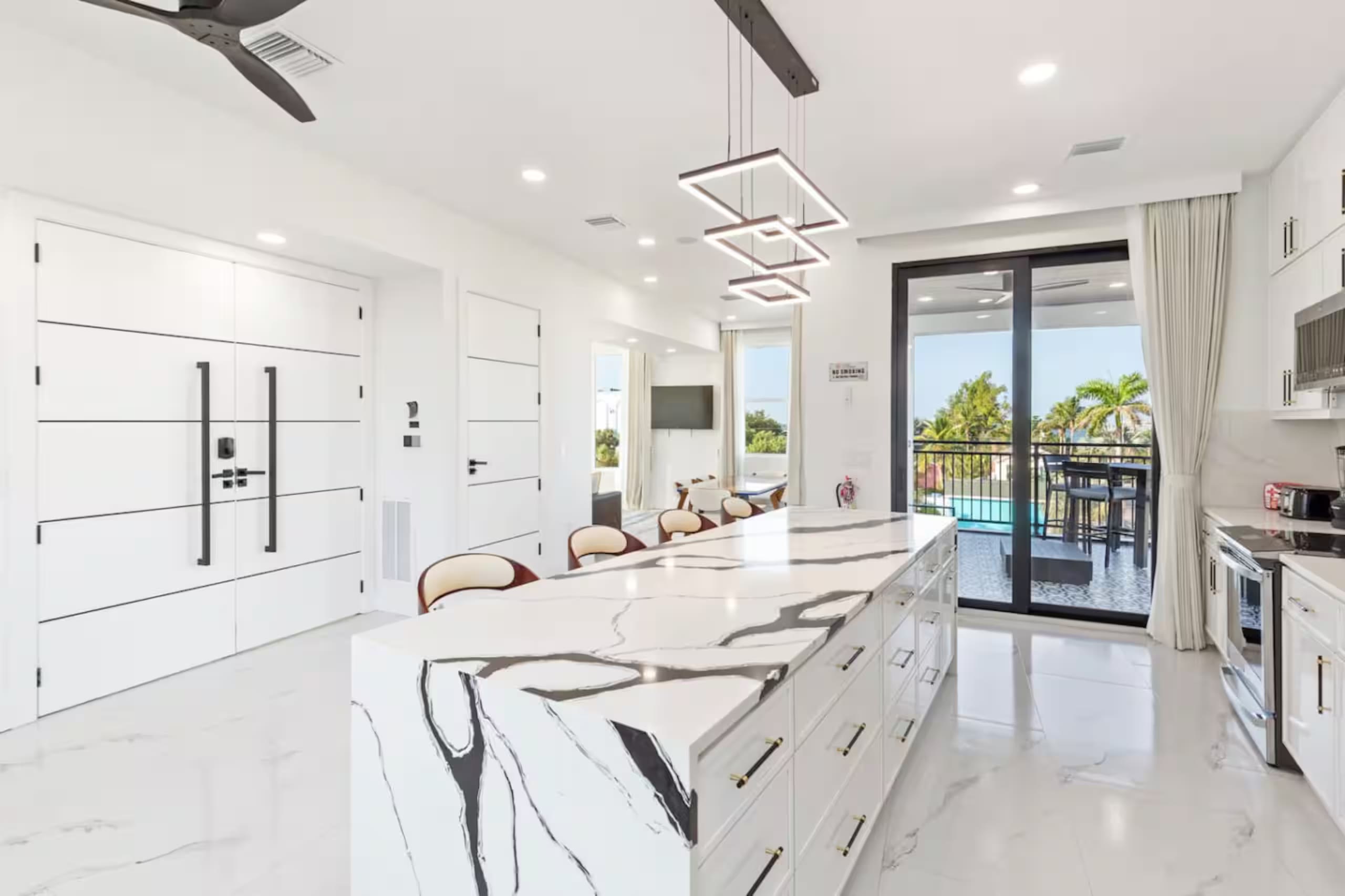 White kitchen with a dark-veined marble waterfall island, pendant lighting, and a floor-to-ceiling glass slider open to a balcony with pool views below