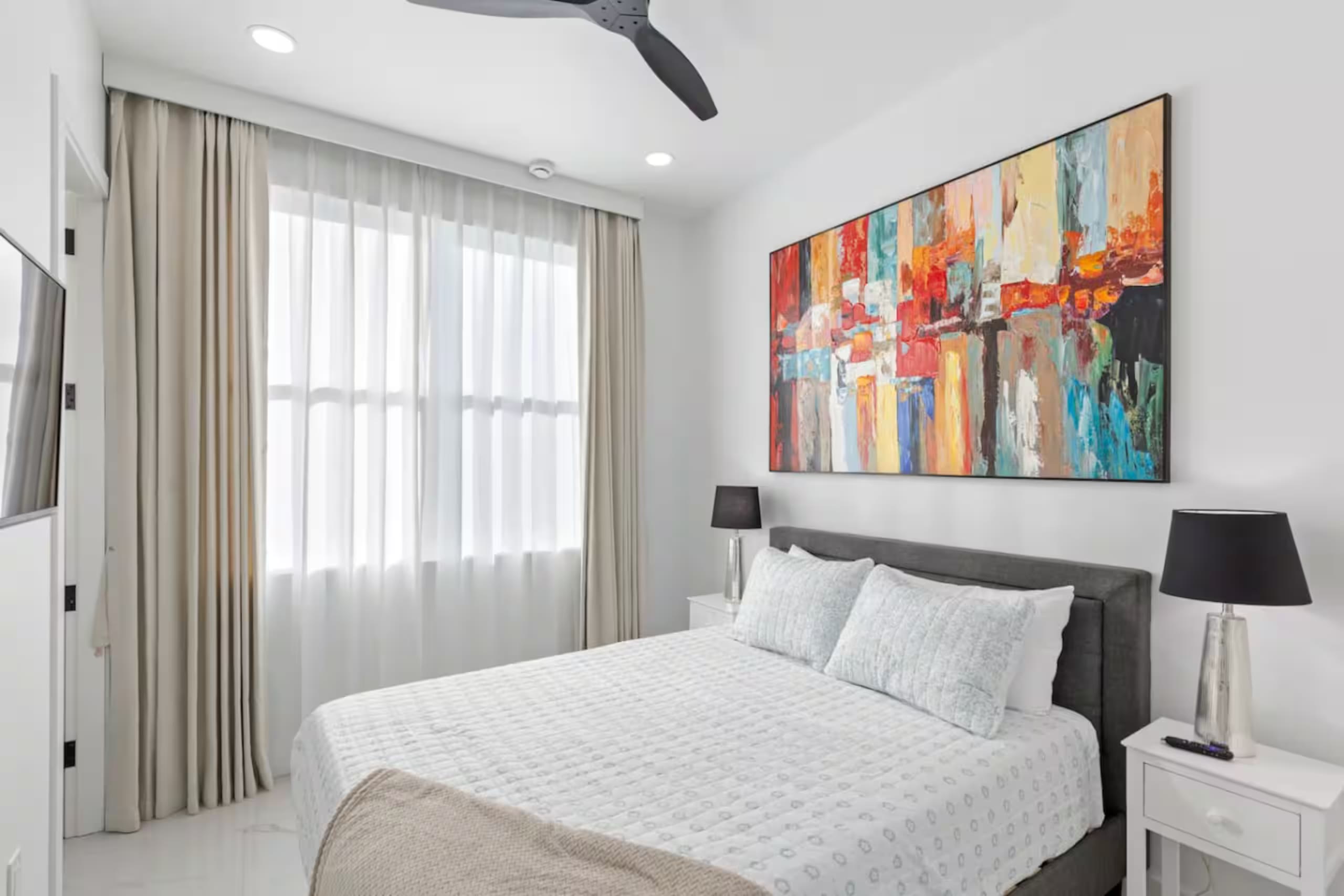 Bedroom with gray upholstered bed, white bedding, nightstand lamps, and a large abstract painting above the headboard