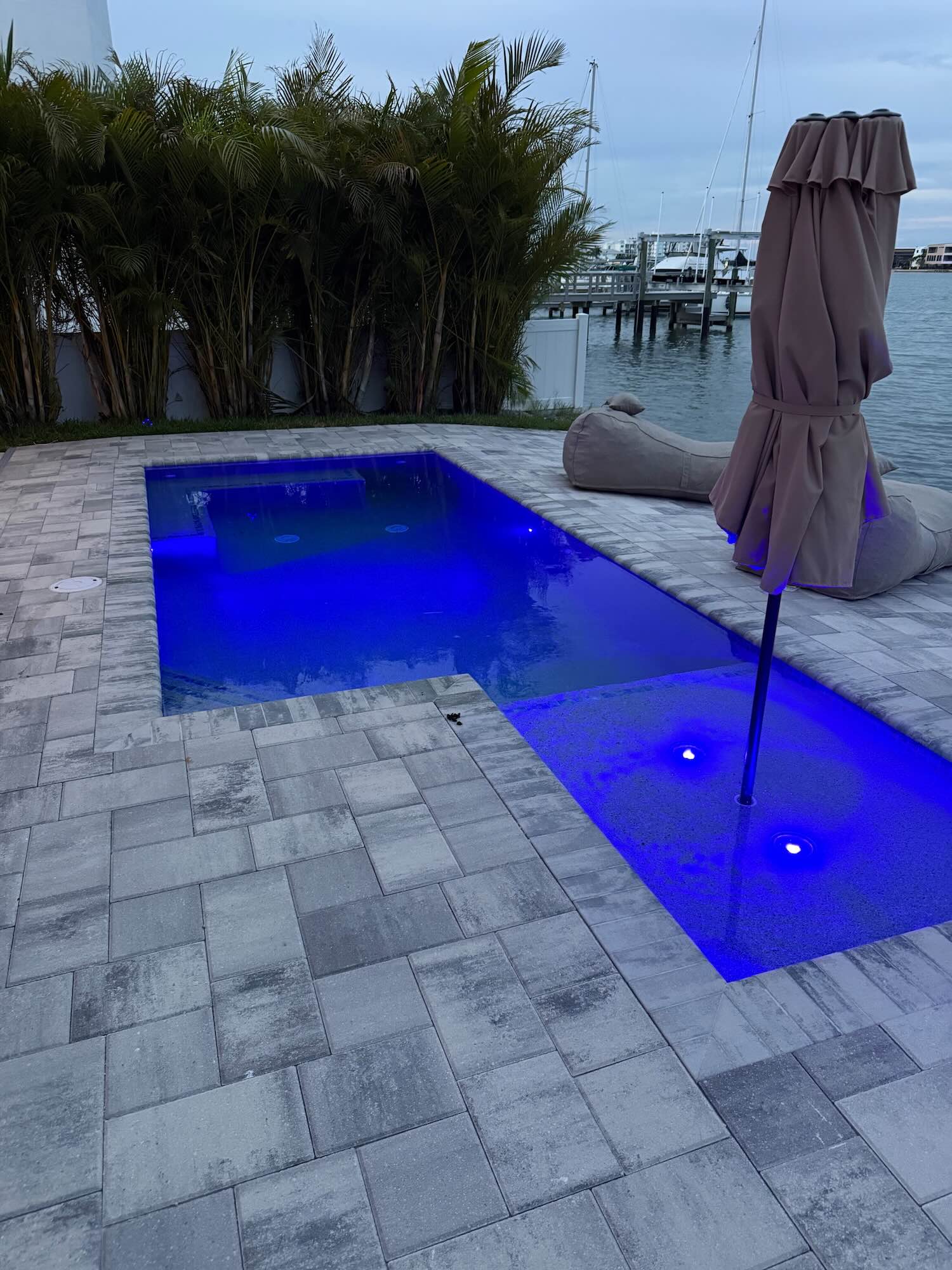 Rectangular pool with an attached raised sun shelf, lit blue at dusk, with a dock and boats visible across the water