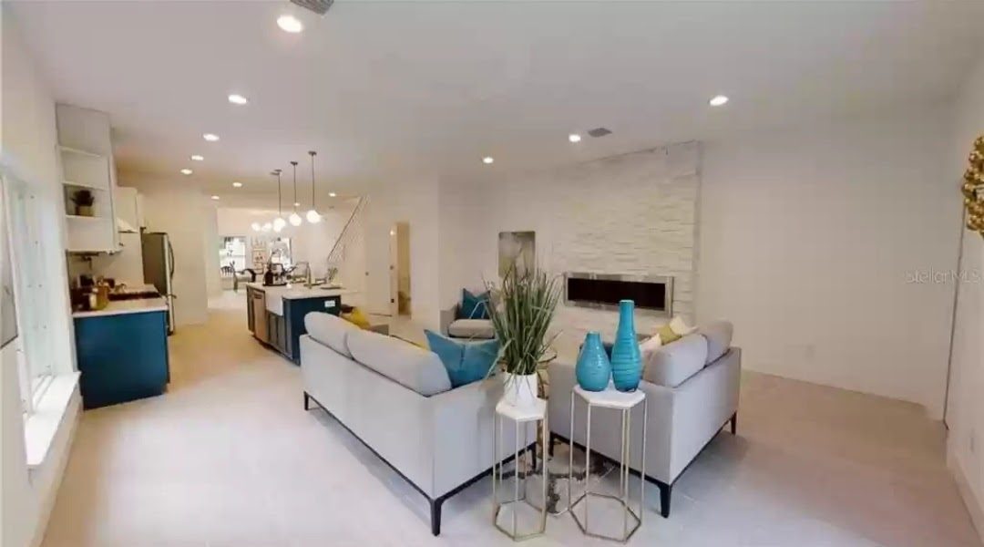 Open-concept living room with gray sectional sofa and stacked-stone linear fireplace, kitchen visible beyond