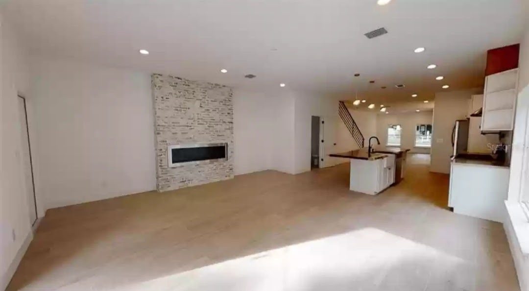 Open living area with stacked-stone fireplace wall and wood-look flooring, kitchen visible in background